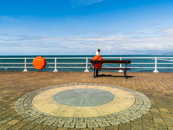 Decorative photo of woman sitting on a bench on a promenade by the sea on a bright sunny day.